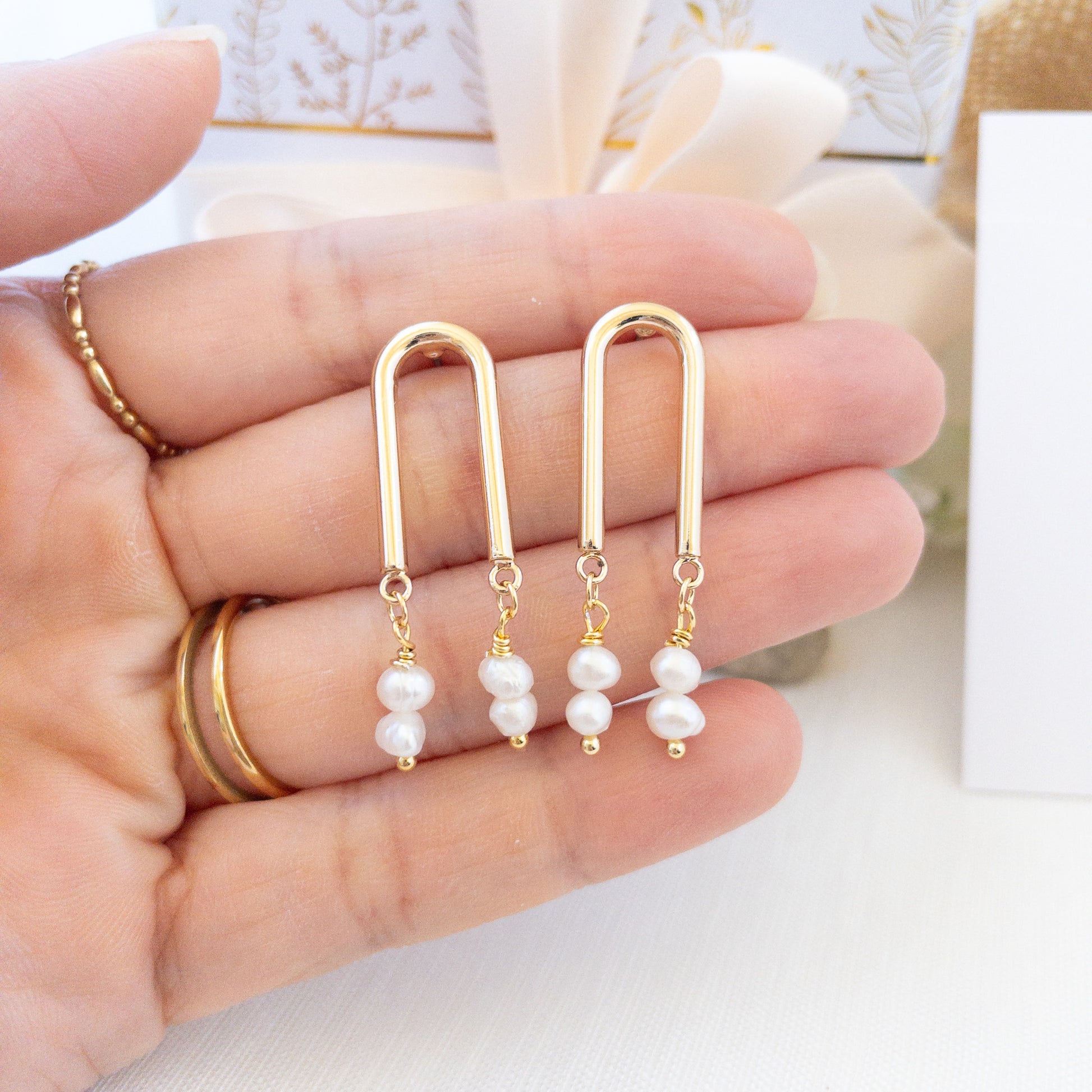 A hand holding a pair of gold arch shaped post earrings with freshwater pearls