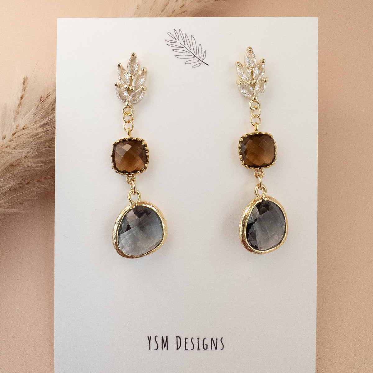 Amber brown and smoky gray crystal drop earrings in warm sunlight on blush background